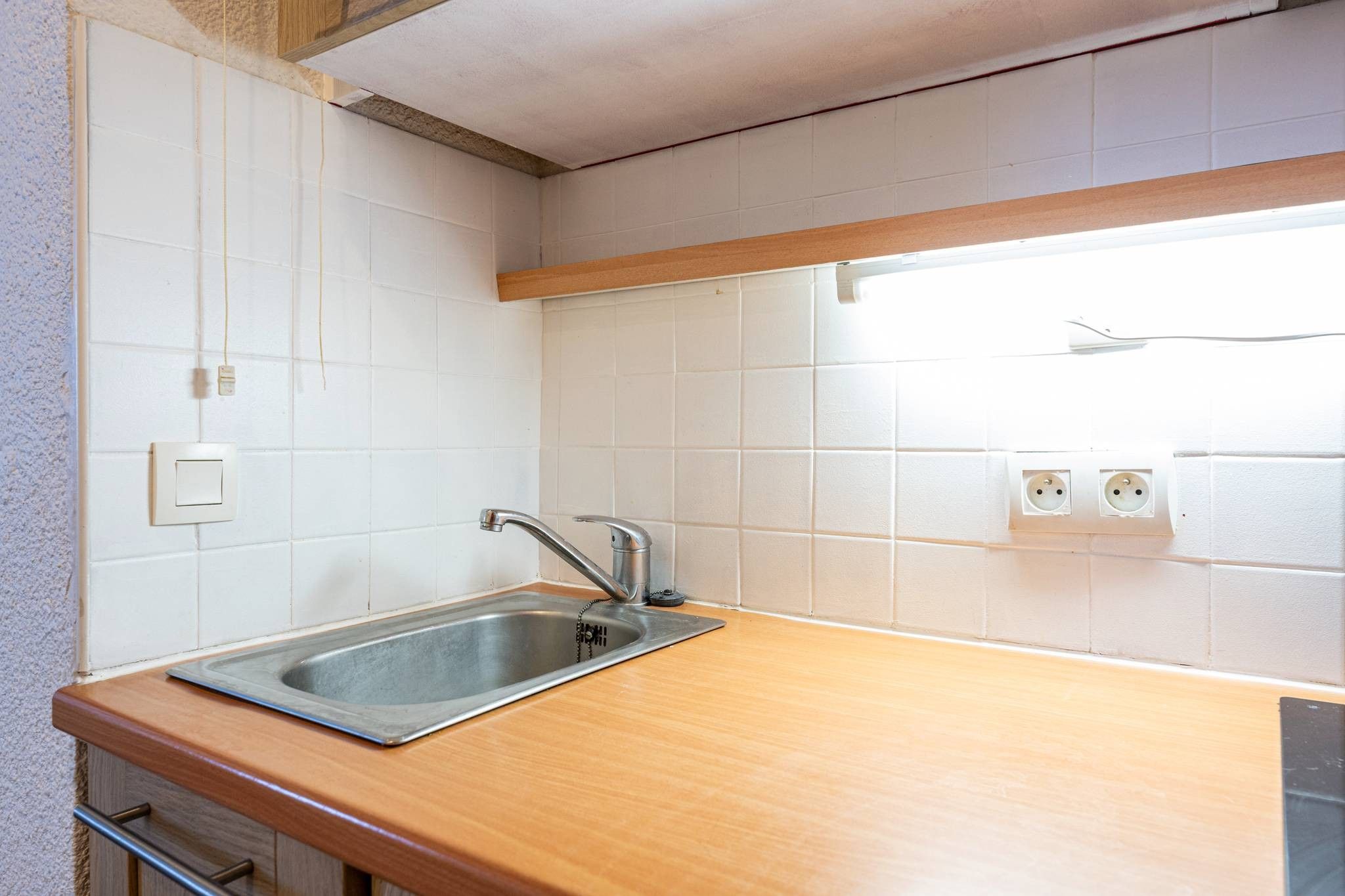 A kitchen with a stainless steel sink, wooden countertop, white tiled backsplash, and electrical outlets.