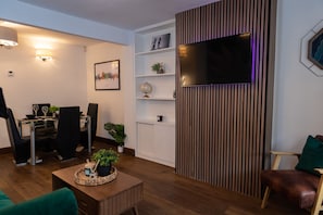 A modern living room with a flat-screen TV mounted on a wooden panel wall, a dining area with a glass table and chairs, and a coffee table with decorative items. - Cosy Windsor Family Escape – Near LEGOLAND, Windsor Castle & London (Windsor)