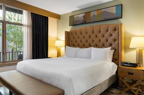 Une chambre d’hôtel avec un grand lit, deux lampes de chevet, un banc, une fenêtre avec des rideaux et un tableau au mur.