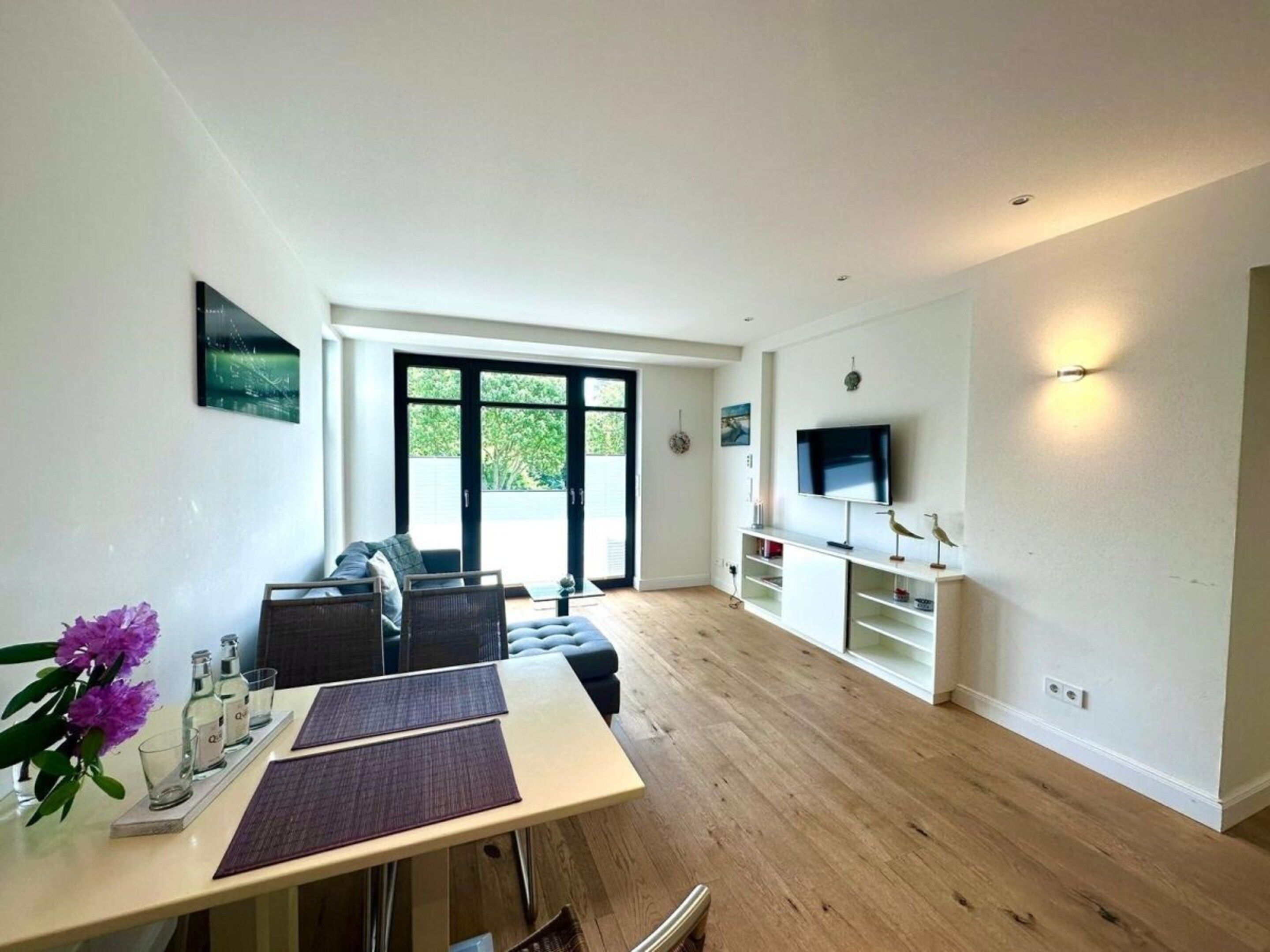 A modern living room with a dining area, large windows, and a flat-screen TV mounted on the wall.