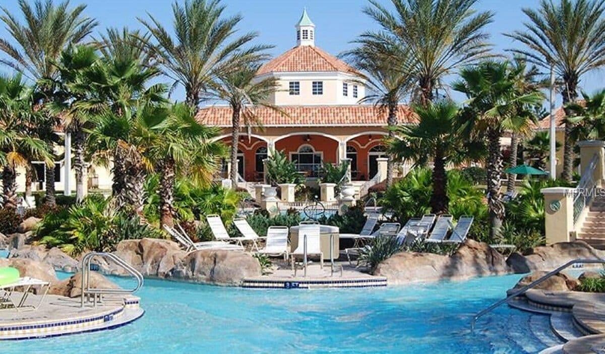 Zona de piscina con sillas de descanso, palmeras y un edificio con techo de tejas rojas.