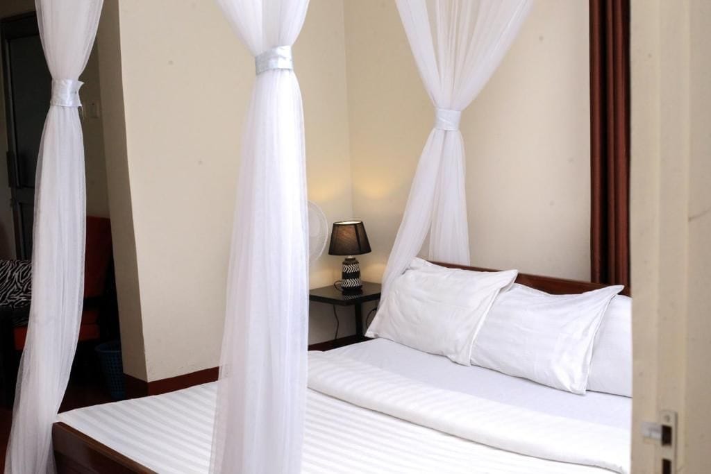 A hotel room with a bed, white curtains, a nightstand with a lamp, and a view of the outside.