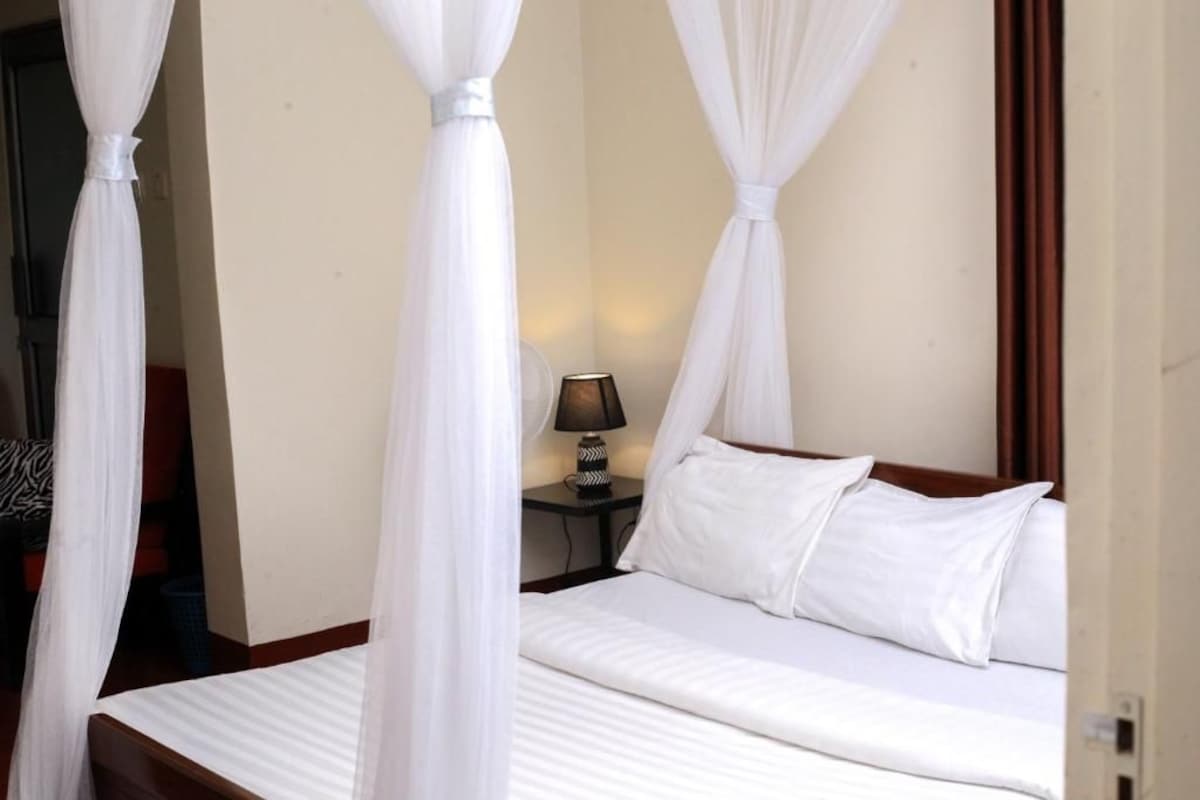 Habitación de hotel con cama, cortinas blancas, mesita de noche con lámpara y vistas al exterior.