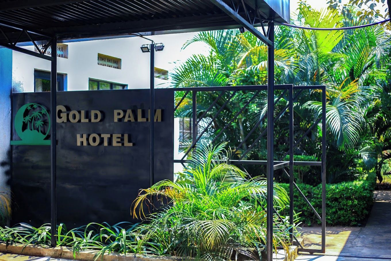 The exterior of the Gold Palm Hotel with a signboard, surrounded by lush greenery and a covered walkway.