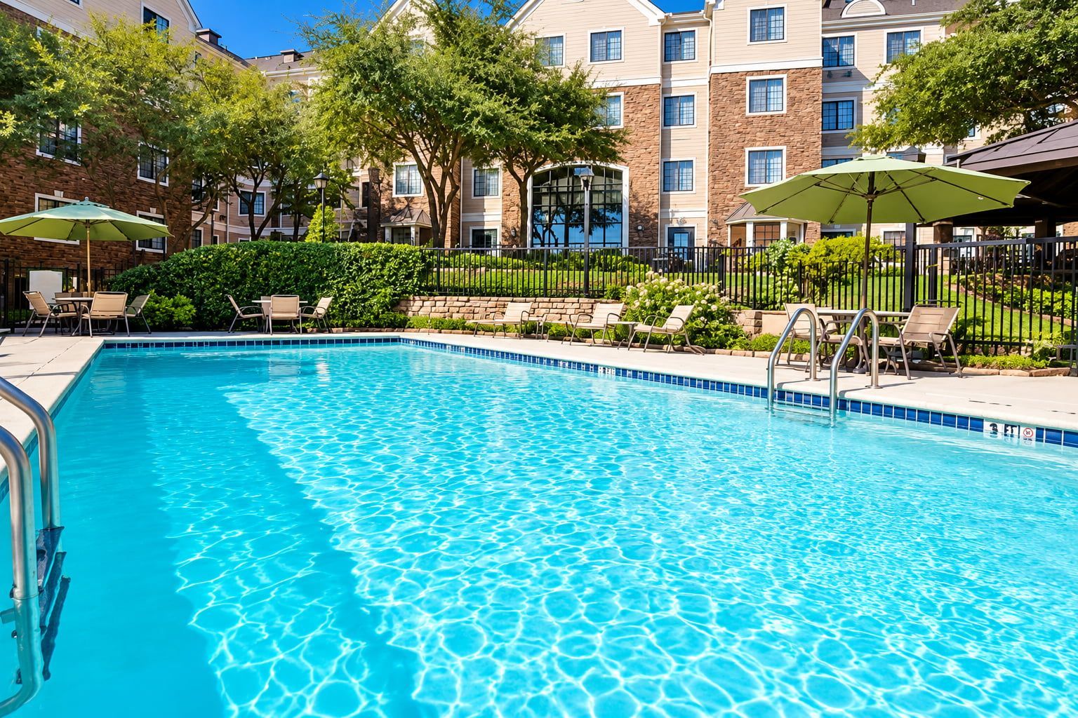 A clear blue pool with lounge chairs and umbrellas, surrounded by a brick building with multiple windows.