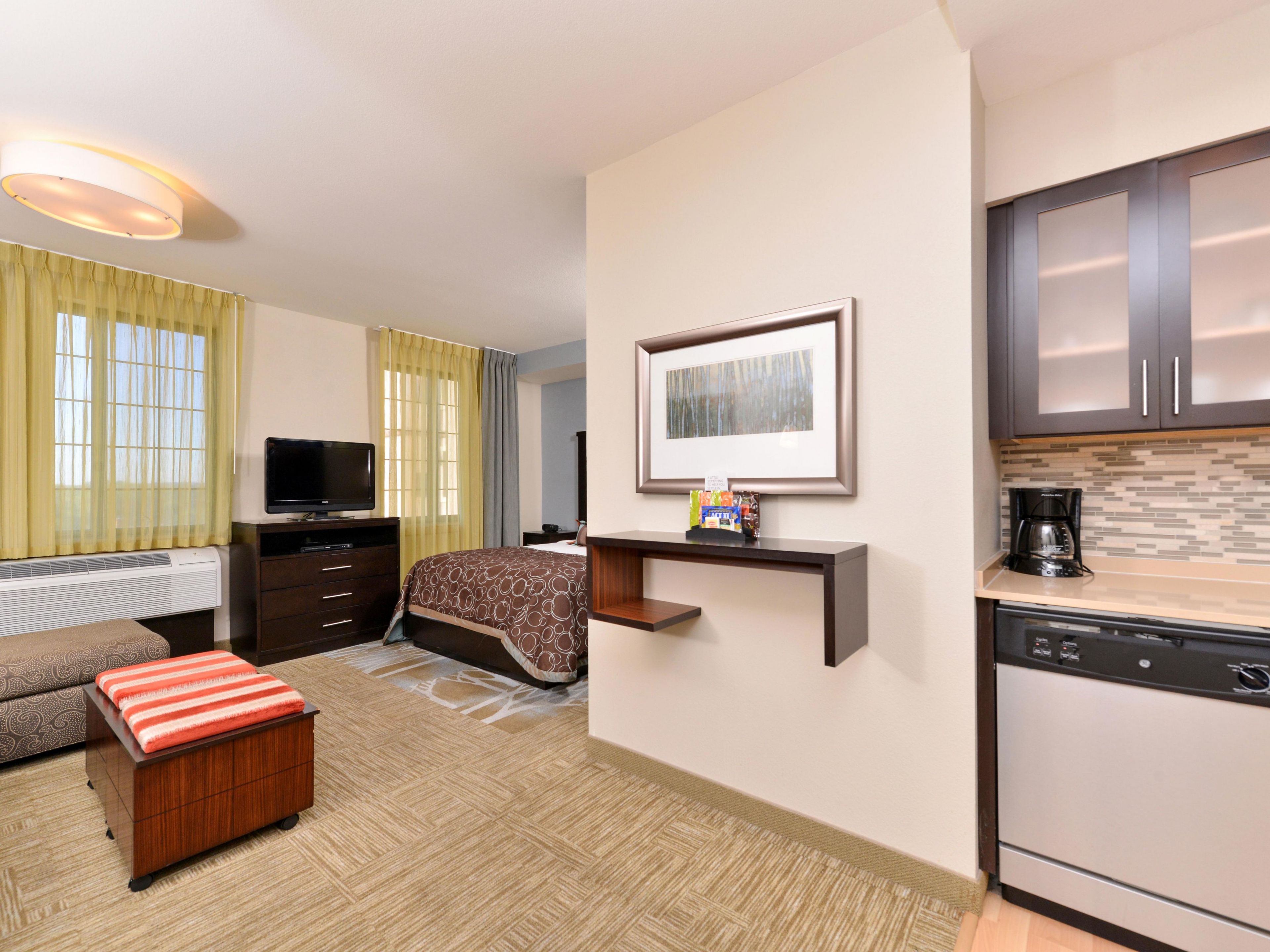 A hotel room with a bed, a television, a small kitchenette, and a framed picture on the wall.