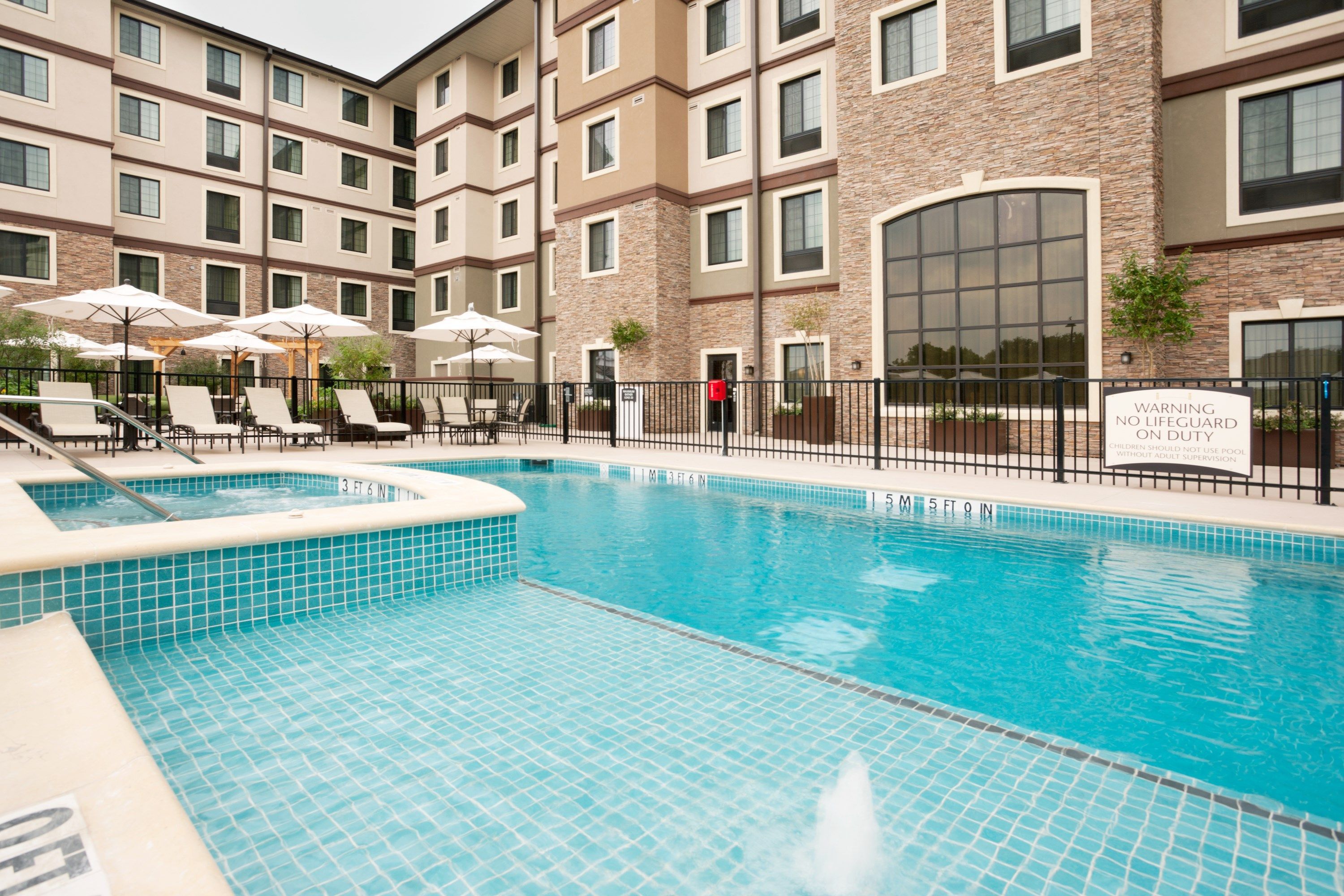 A swimming pool with lounge chairs and umbrellas, surrounded by a building with multiple windows and a sign warning about no lifeguard on duty.