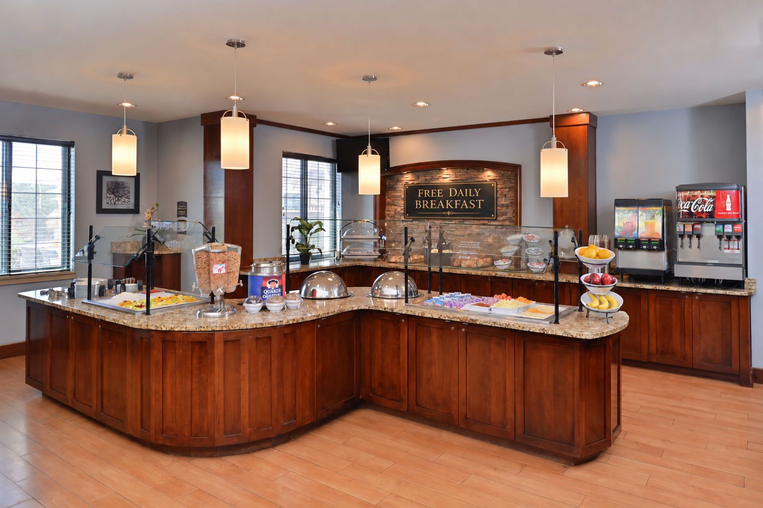 A breakfast buffet counter with a sign indicating free daily breakfast, various food items, and a Coca-Cola dispenser.