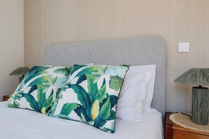 Un lit avec une tête de lit grise, deux oreillers ornés d’un motif de feuilles tropicales et une lampe de chevet posée sur une table de nuit en bois.