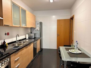 A compact kitchen with a stove, sink, and refrigerator. There is a small dining table with a water bottle and a glass. - Sea view - Peaceful Atlantic View by Cristo Rei (Caniço)