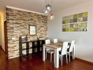 A dining area with a stone wall, a wooden table, white chairs, and hanging pendant lights. - Sea view - Peaceful Atlantic View by Cristo Rei (Caniço)
