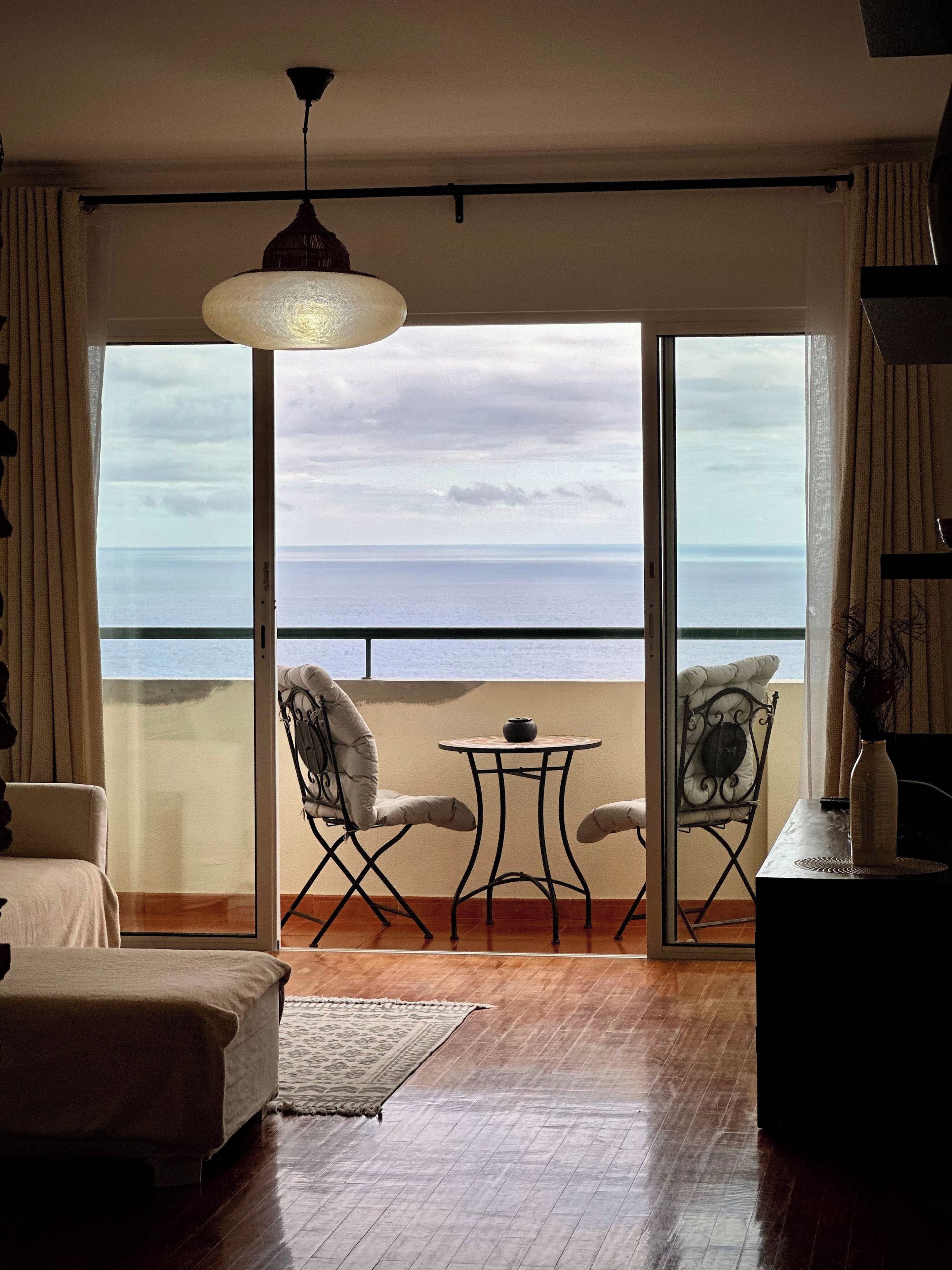 A room with a view of the sea through large glass doors, a round table with chairs, and a hanging light fixture.