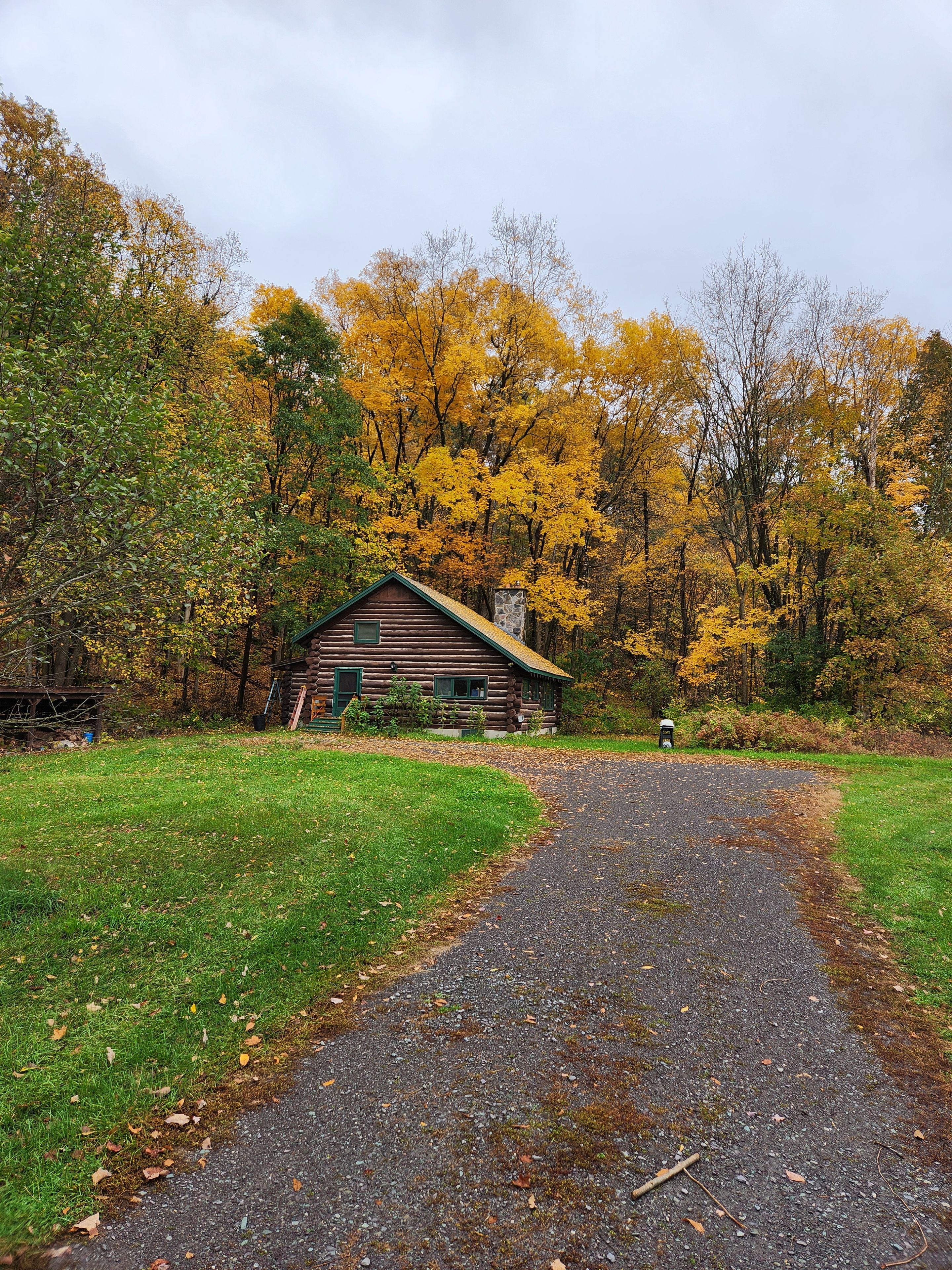 A rustic cabin surrounded by autumn foliage, a gravel path leading to it, and a stone structure in the background.