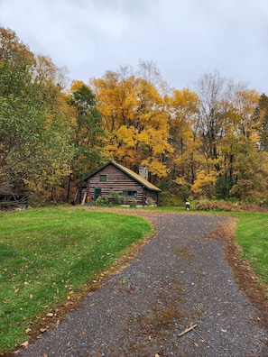 A rustic cabin surrounded by autumn foliage, a gravel path leading to it, and a stone structure in the background.