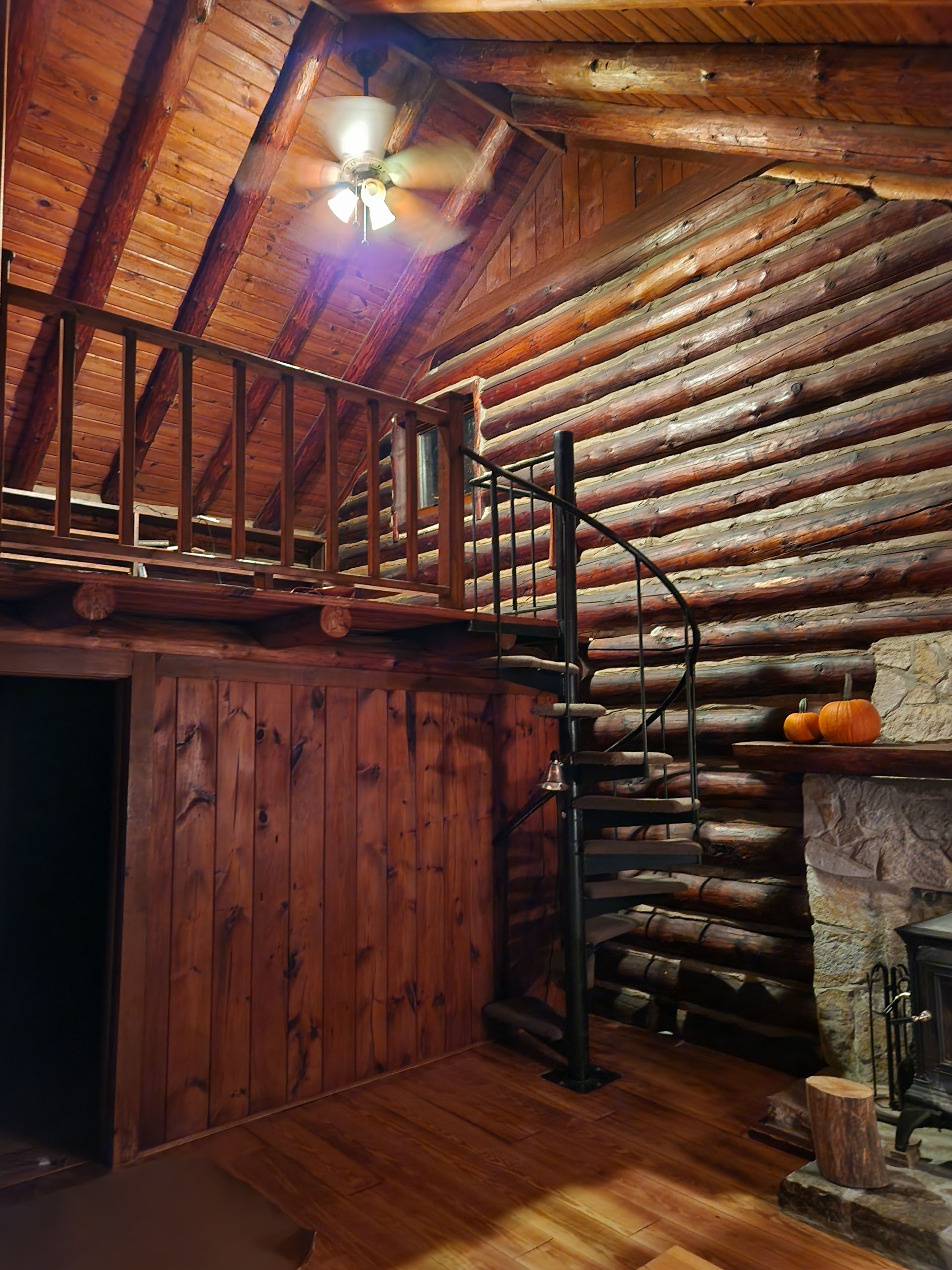 A log cabin interior with a wooden staircase, a stone fireplace, and a ceiling fan.