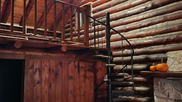 A log cabin interior with a wooden staircase, a stone fireplace, and a ceiling fan.