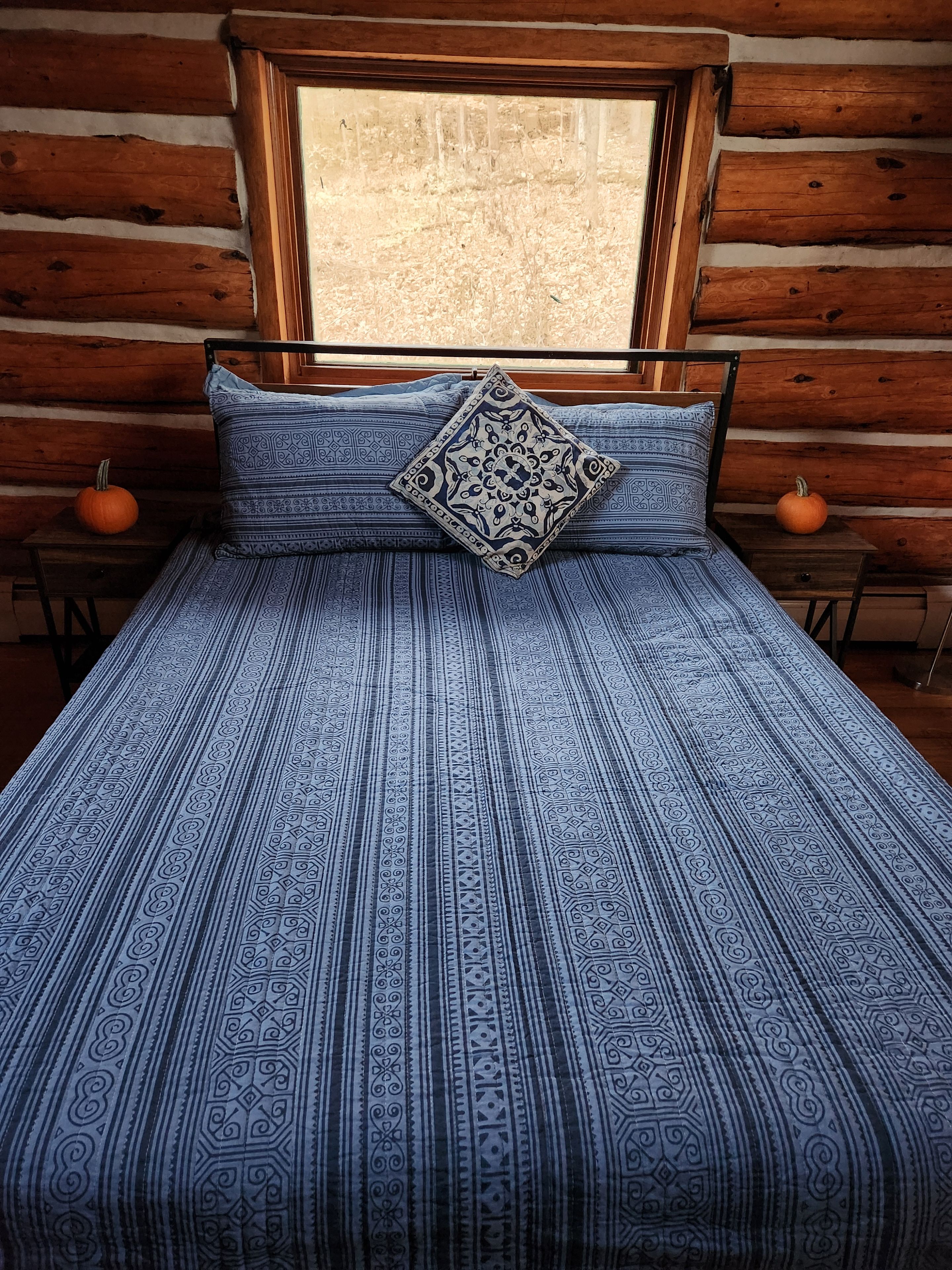 A bed with a patterned blue and white bedspread, a decorative pillow, and two pumpkins on wooden nightstands.
