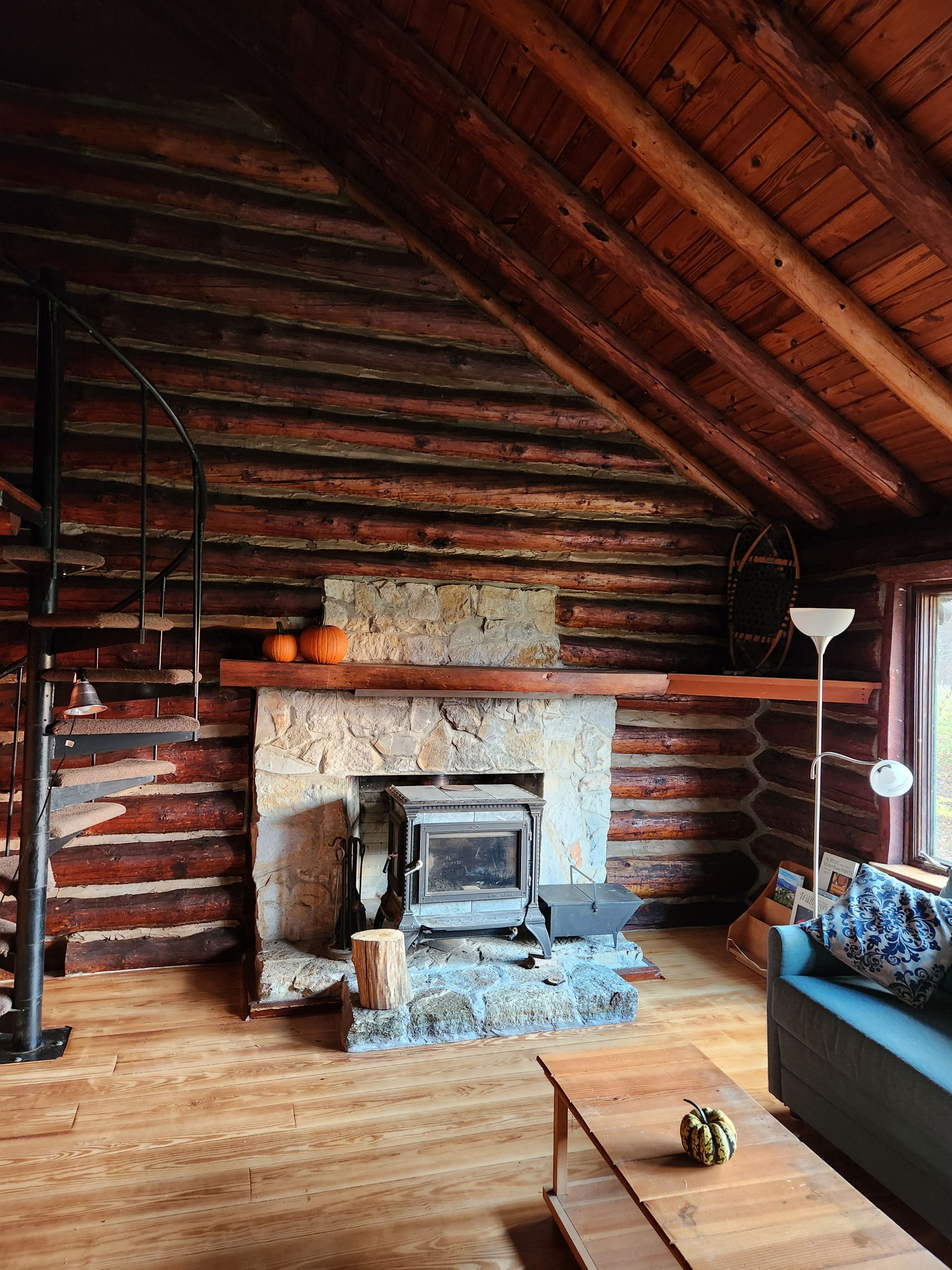 Interior rústico de una cabaña con chimenea de piedra, pisos de madera, una escalera en espiral y una zona de estar acogedora.