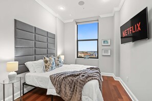 A modern bedroom with a large bed, a grey headboard, a window with a city view, a mounted TV, and a bedside table with a lamp. - Charming NoLib Studio | Sleeps 2 | Onsite Fitness (Philadelphia)