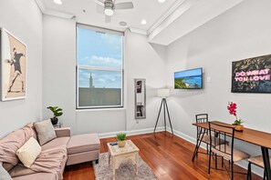 A modern living room with a large window, a sofa, a coffee table, a dining table with chairs, and a floor lamp. - Vibrant Home 1BD | Quick Transit Sights | Stay 2 (Philadelphia)