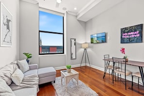 A modern living room with a grey sofa, a small white table, a dining area with a wooden table and chairs, a floor lamp, and a large window with a view of a building and sky. - Vibrant Home 1BD | Quick Transit Sights | Stay 2 (Philadelphia)