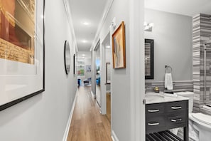 Un couloir doté d’un plancher en bois, d’un mur blanc orné d’un tableau encadré, et d’une salle de bain avec un lavabo et un miroir modernes.