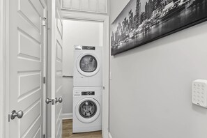 Un lavadero con dos lavadoras y secadoras apiladas, una puerta con pomo redondo y un cuadro de paisaje urbano en la pared.