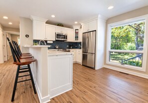 Une cuisine moderne avec des meubles blancs, des appareils en acier inoxydable et une grande fenêtre donnant sur un paysage verdoyant.