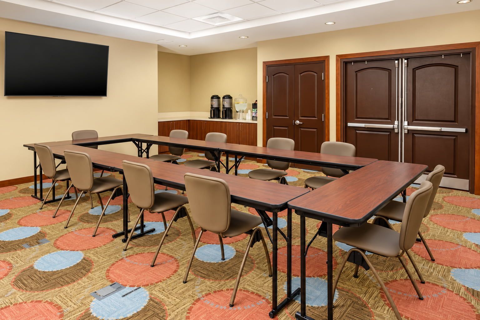 Une salle de conférence équipée d’une longue table, de chaises, d’un téléviseur à écran plat, d’un coin café et de portes à deux battants.