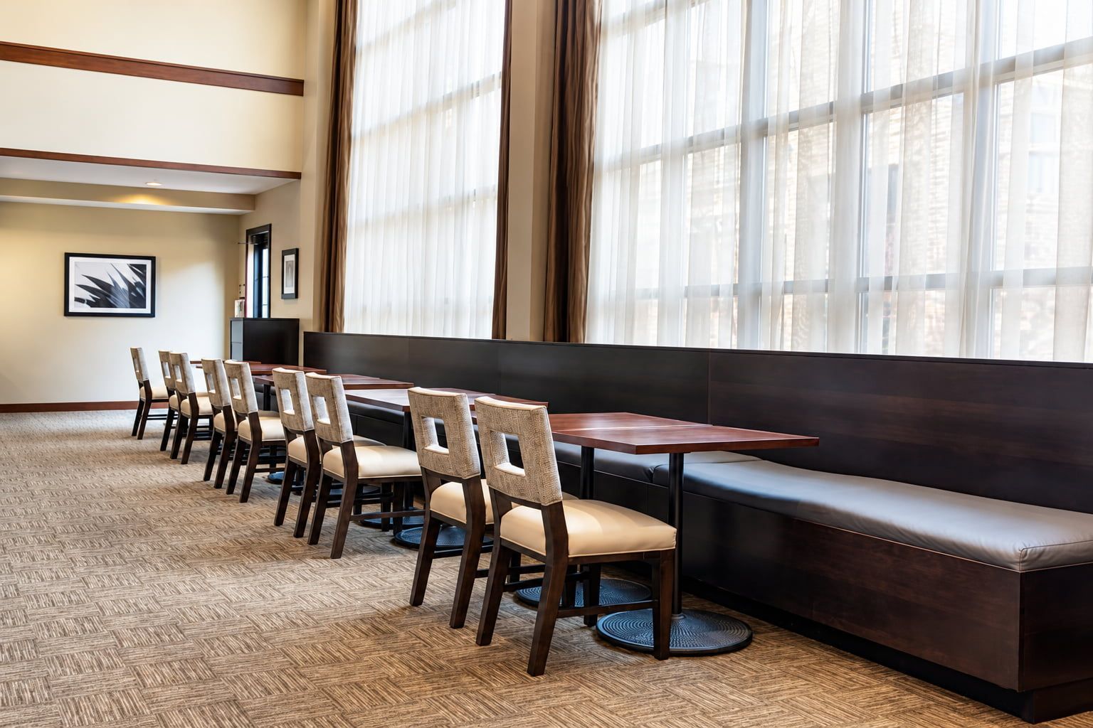 Une salle à manger d’hôtel avec une longue table, six chaises, un banc et de grandes fenêtres ornées de rideaux légers.