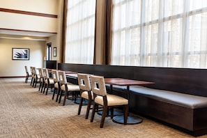 Une salle à manger d’hôtel avec une longue table, six chaises, un banc et de grandes fenêtres ornées de rideaux légers.