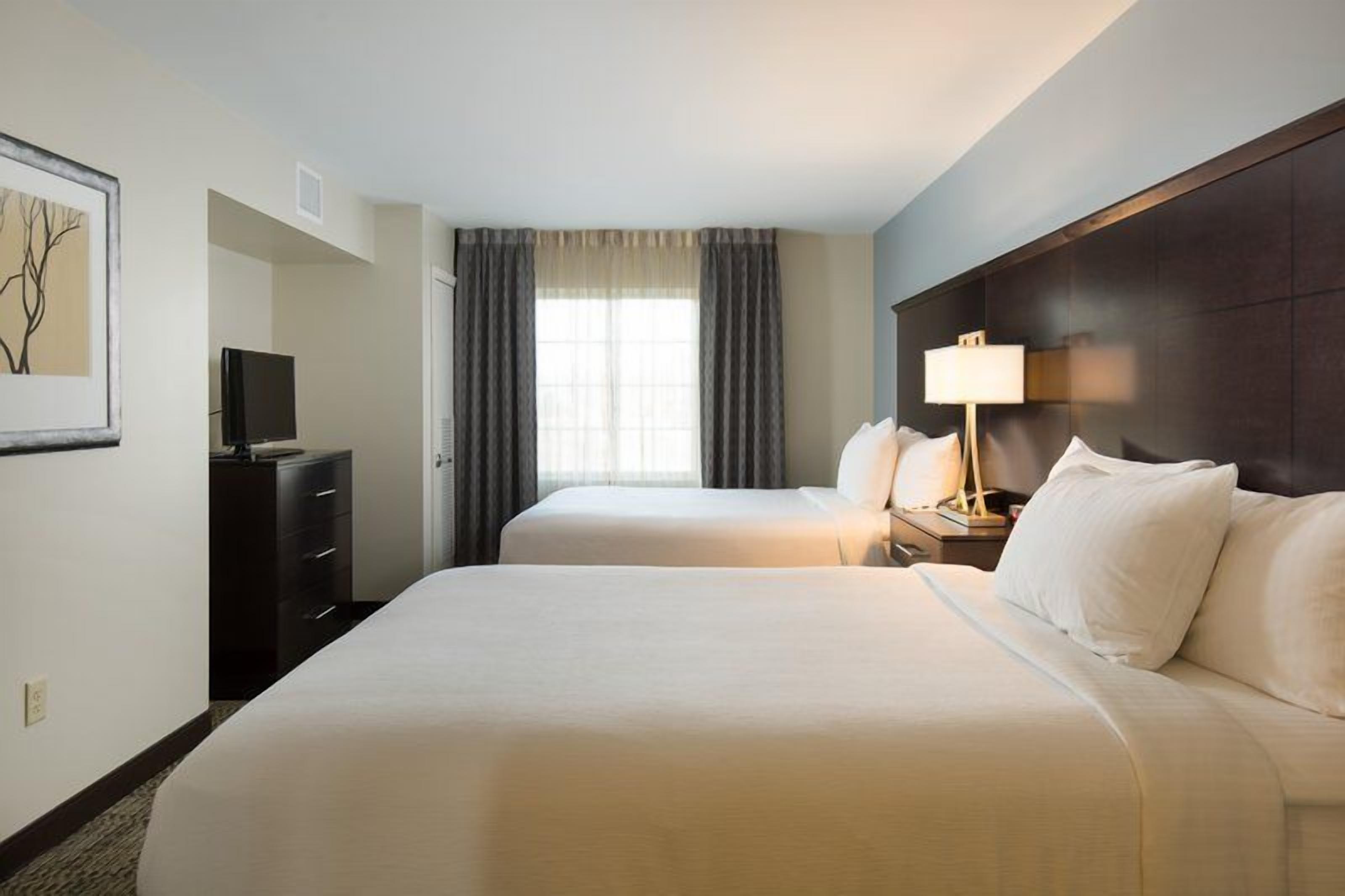 Une chambre d’hôtel avec deux lits, un téléviseur posé sur une commode, une table de chevet avec une lampe et une fenêtre avec des rideaux.