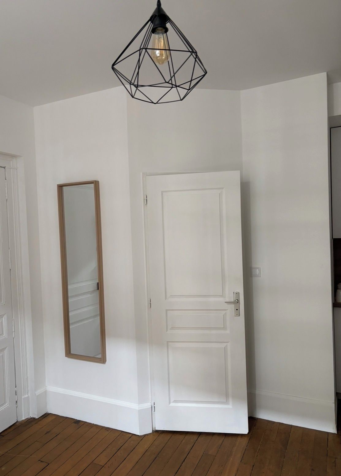 Quarto com porta branca, piso de madeira e luminária pendente geométrica.