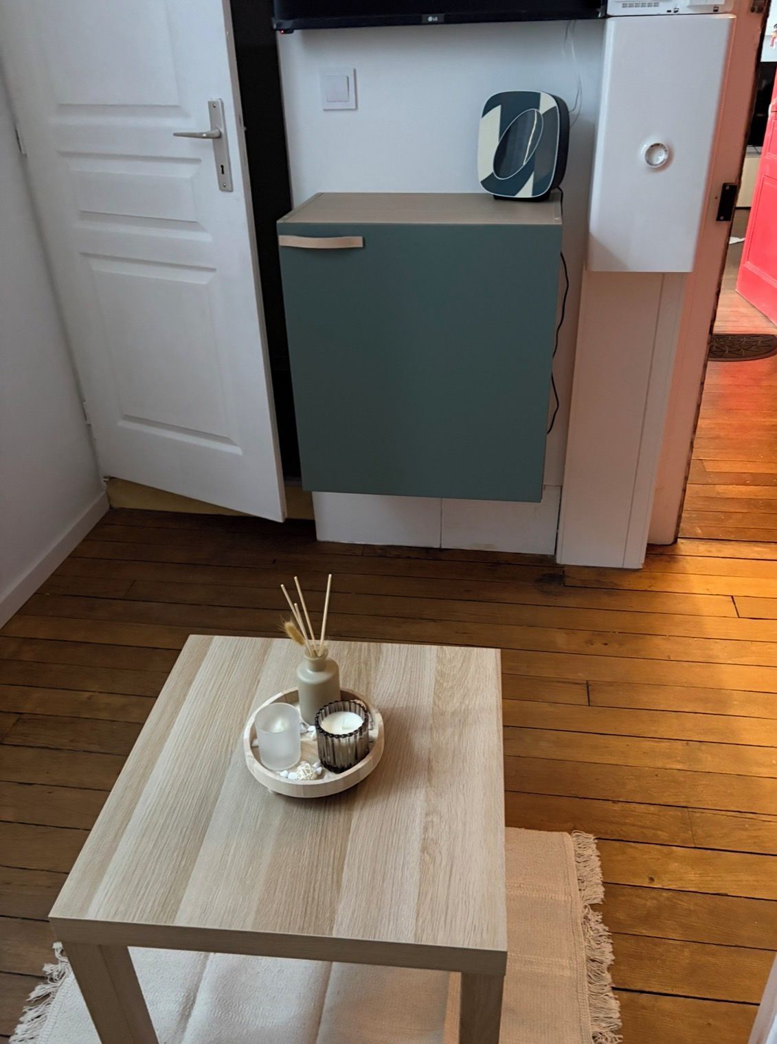 Uma pequena mesa de madeira com uma vela e um vaso pequeno, uma porta branca e uma cozinheira com geladeira e micro-ondas.