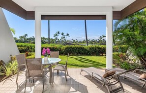 A covered outdoor seating area with a table and chairs, a lounge chair, and a view of a well-maintained garden with palm trees and a hedge. - Mauna Lani Point - 4 BR (2 condos), Sleeps 8 (Waimea)