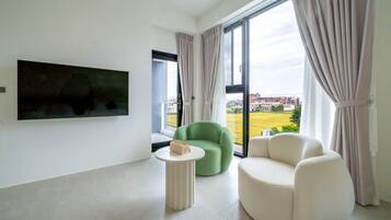 A modern living room with a flat-screen TV mounted on the wall, a green armchair, a white sofa, and a small round table with a handbag on it.