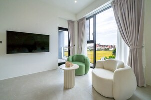 Un salón moderno con un televisor de pantalla plana montado en la pared, un sillón verde, un sofá blanco y una pequeña mesa redonda con un bolso encima.