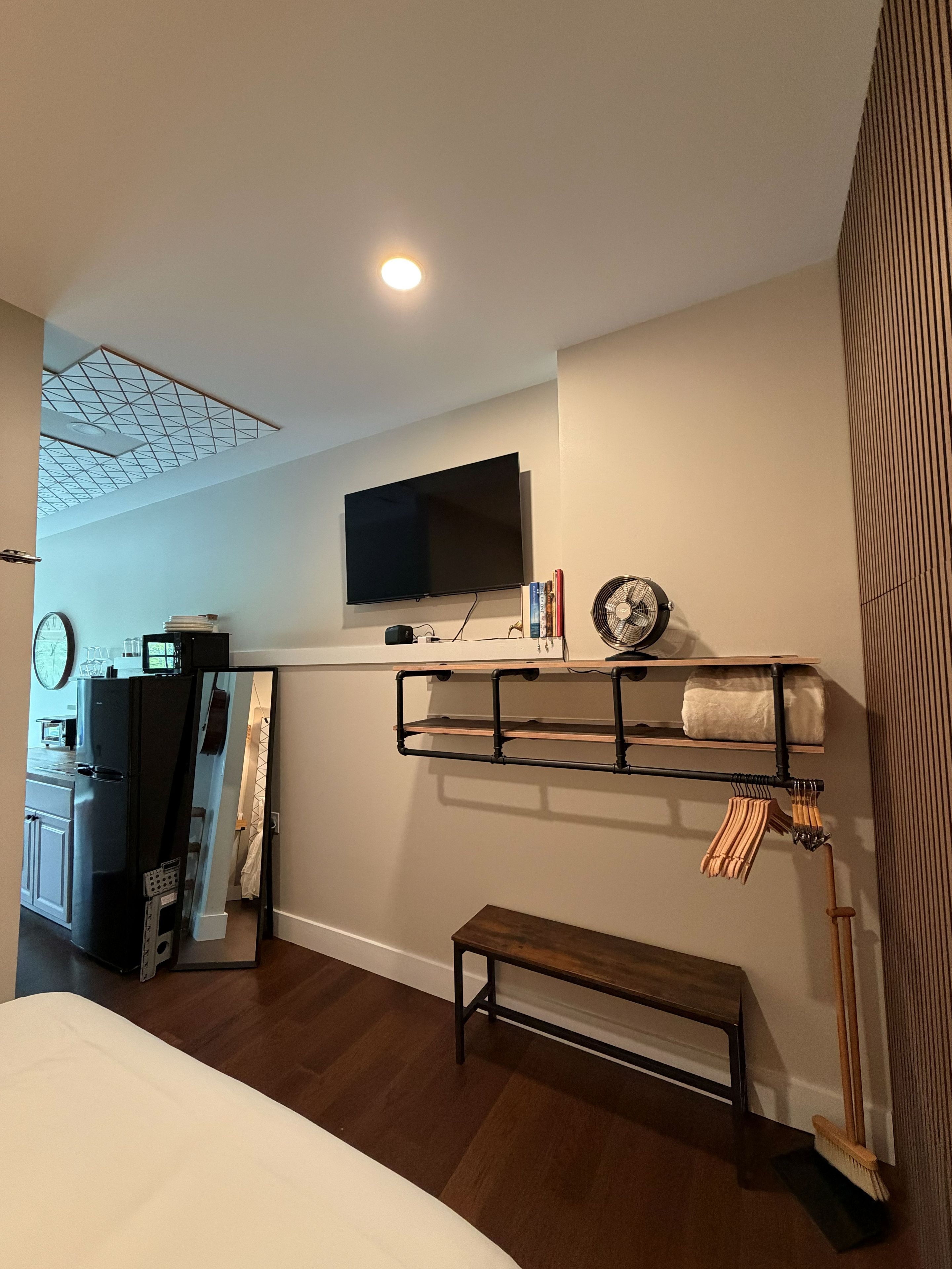 A bedroom with a bed, a TV mounted on the wall, a shelf with books and a fan, and a wooden bench.