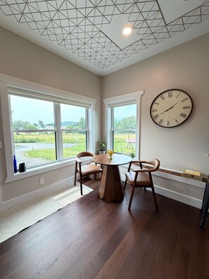 A room with a wooden dining table, two chairs, a wall clock, and large windows with a view of greenery. - Unique Boutique Studio Apartment Getaway for Two. (Underhill)