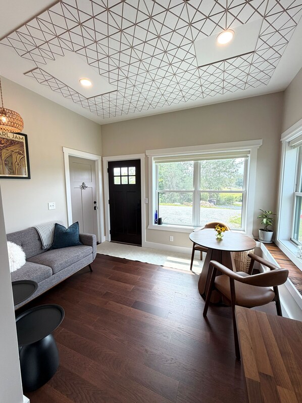 Un salon moderne avec un canapé, une table à manger et des chaises, une porte noire et une fenêtre donnant sur un paysage verdoyant.