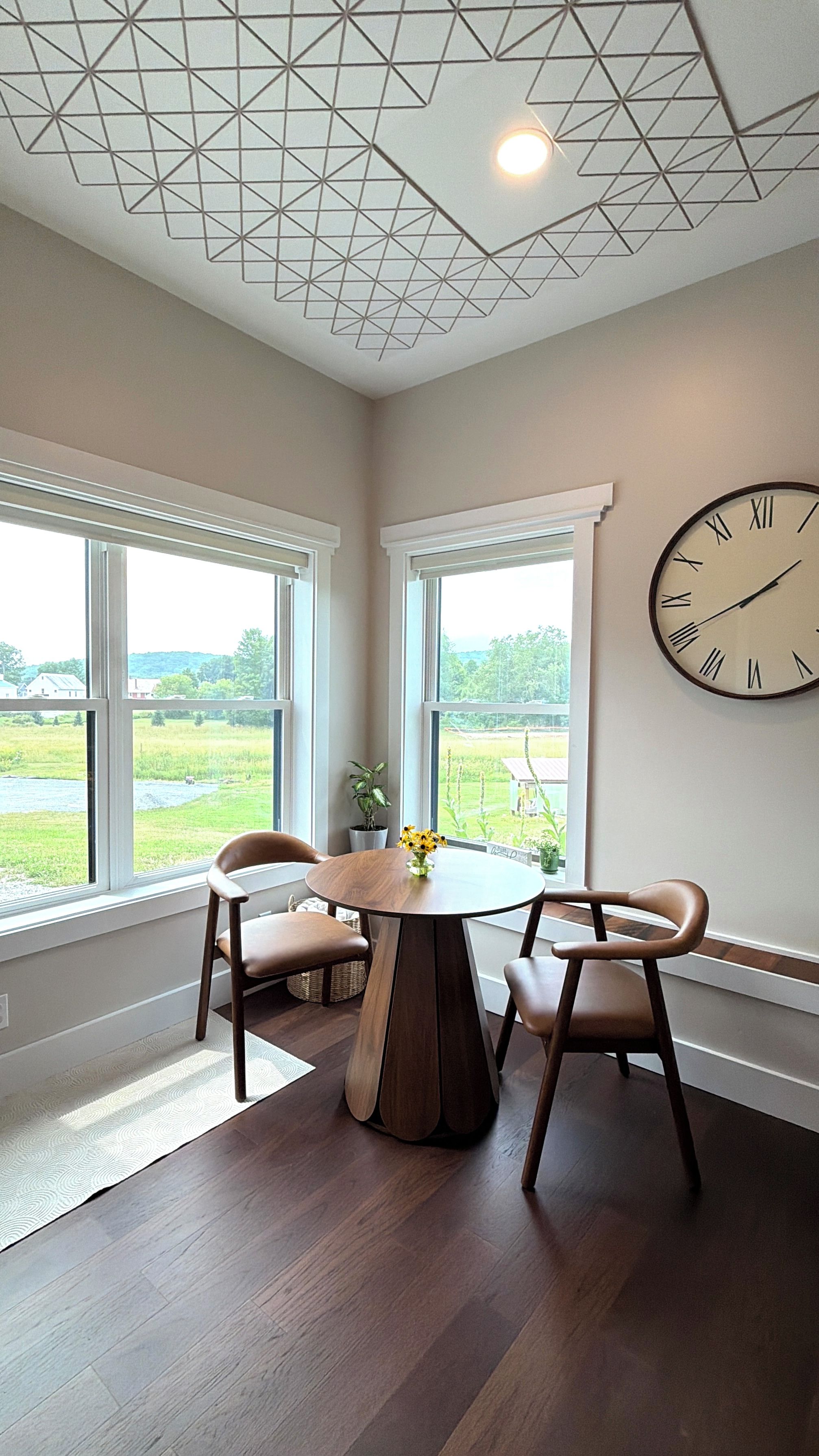 A dining area with a round table, two chairs, a clock on the wall, and large windows with a view of a green landscape.