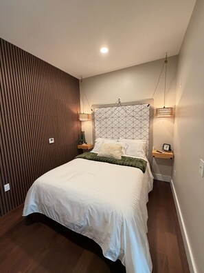 Un lit bien fait, recouvert d’une couette blanche et d’un plaid vert, flanqué de deux tables de chevet avec des lampes, dans une chambre moderne aux murs lambrissés et dotée d’une tête de lit à motifs géométriques.