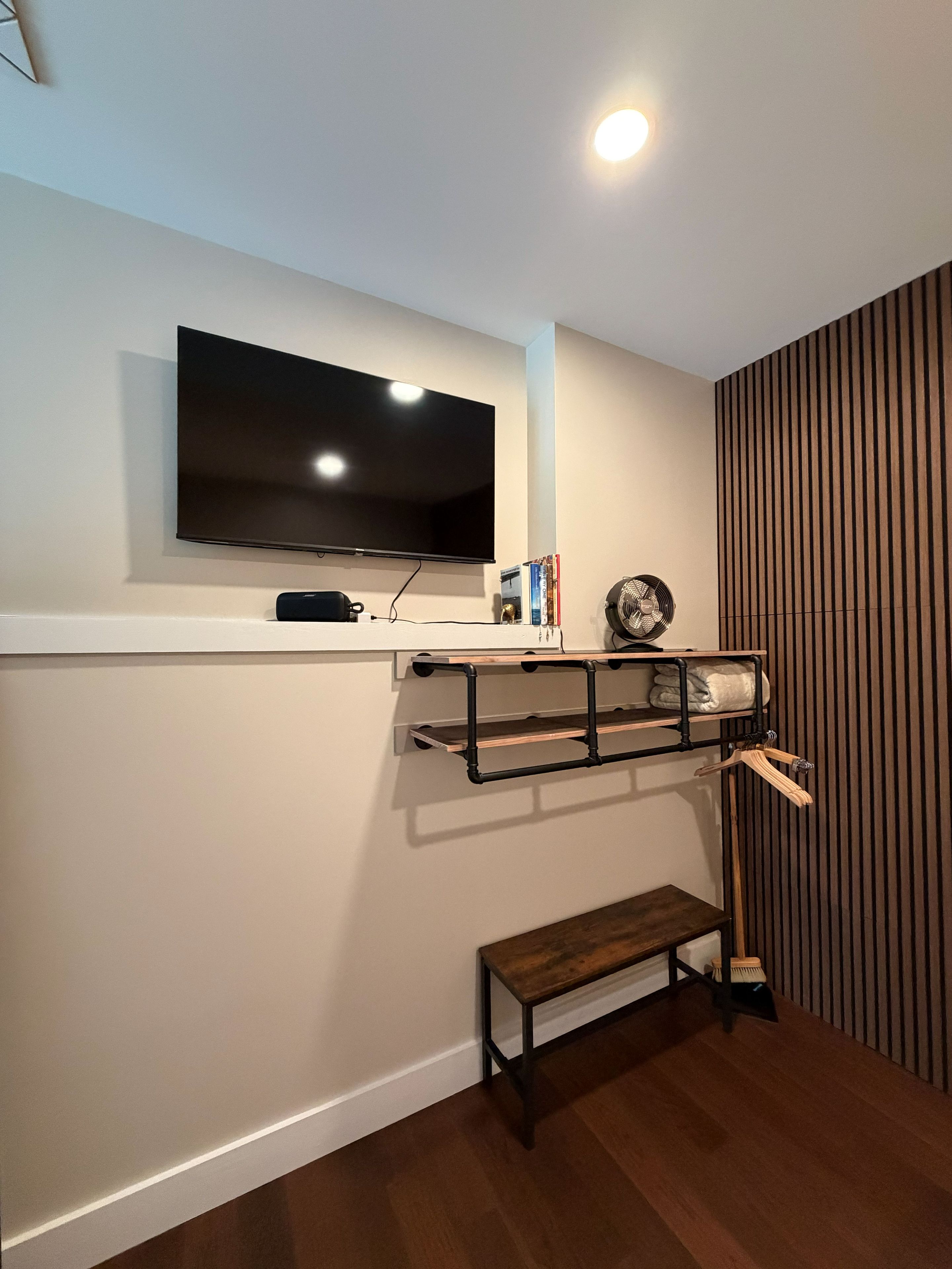 A modern living room with a mounted flat-screen TV, a small wooden bench, and a wall-mounted shelf with books and a fan.