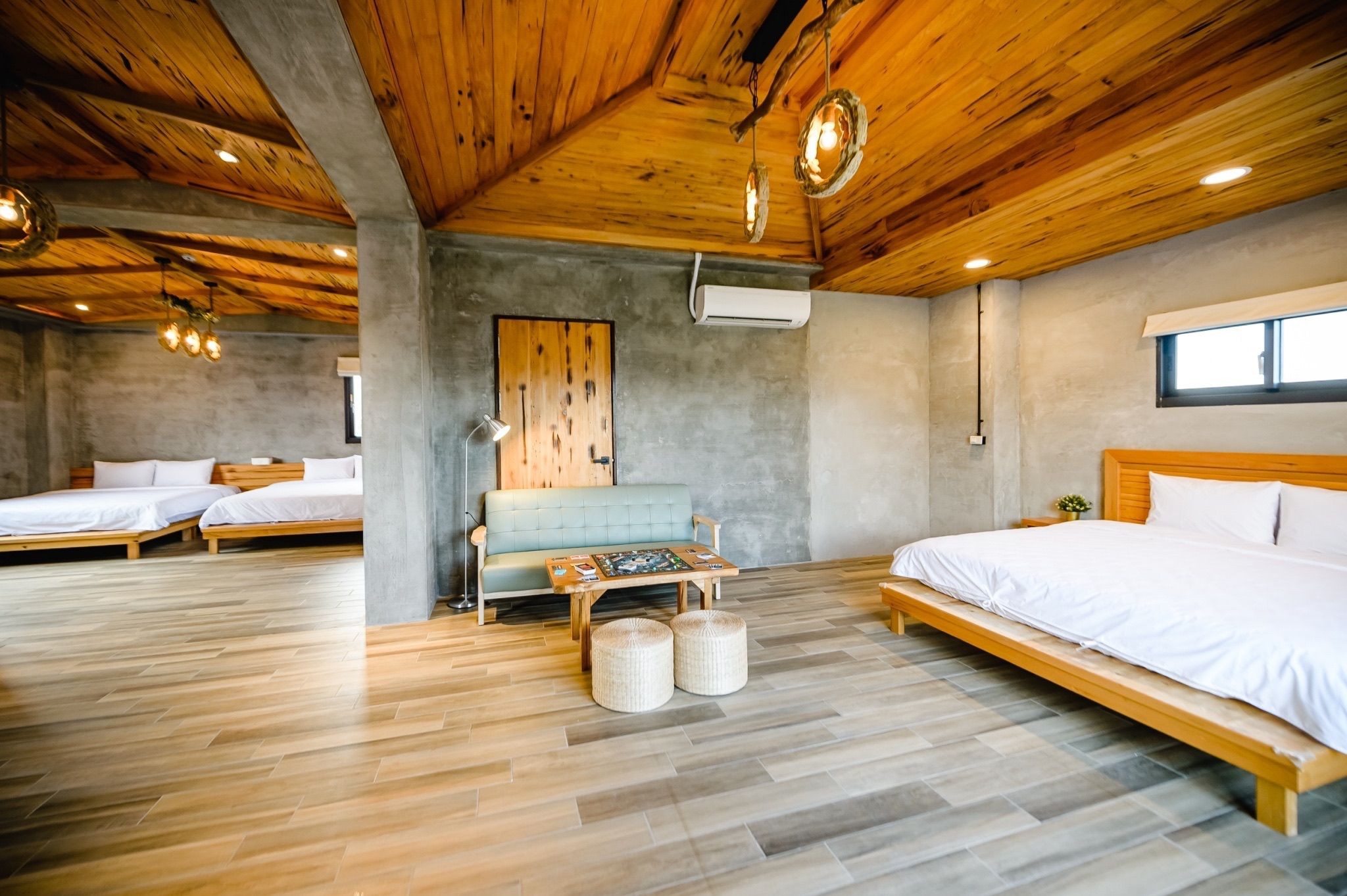 A modern hotel room with a wooden ceiling, a large bed, two smaller beds, a sofa, a table with board games, and a window.