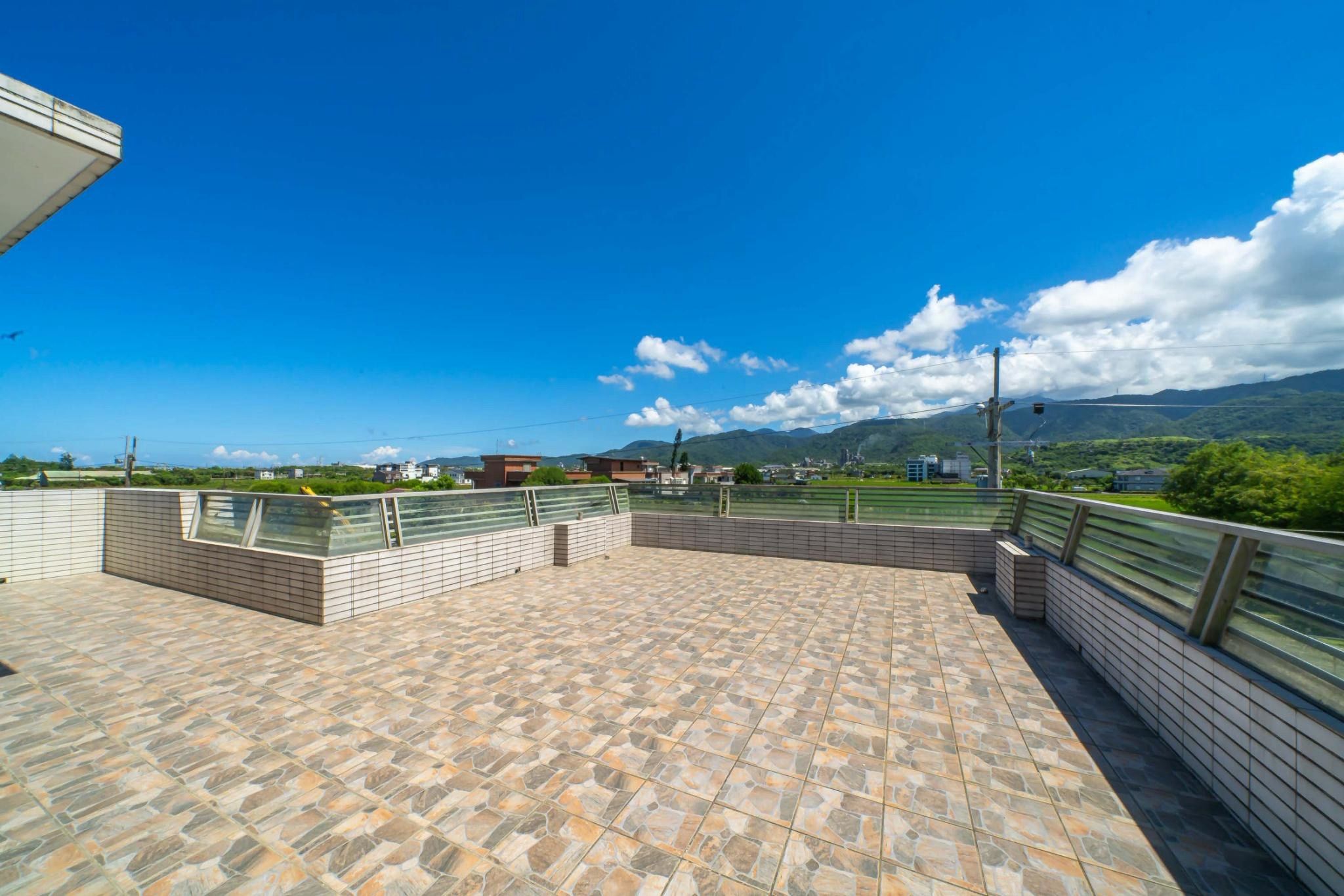 A rooftop terrace with a stone tile floor, metal railings, and a view of distant buildings and mountains.
