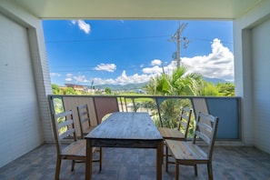 Varanda com mesa e cadeiras de madeira, com vista para uma paisagem montanhosa, vegetação exuberante e céu claro.