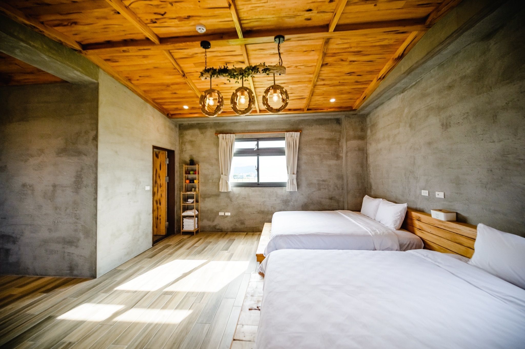 A bedroom with two beds, a wooden ceiling, a window with curtains, and a shelf with items.