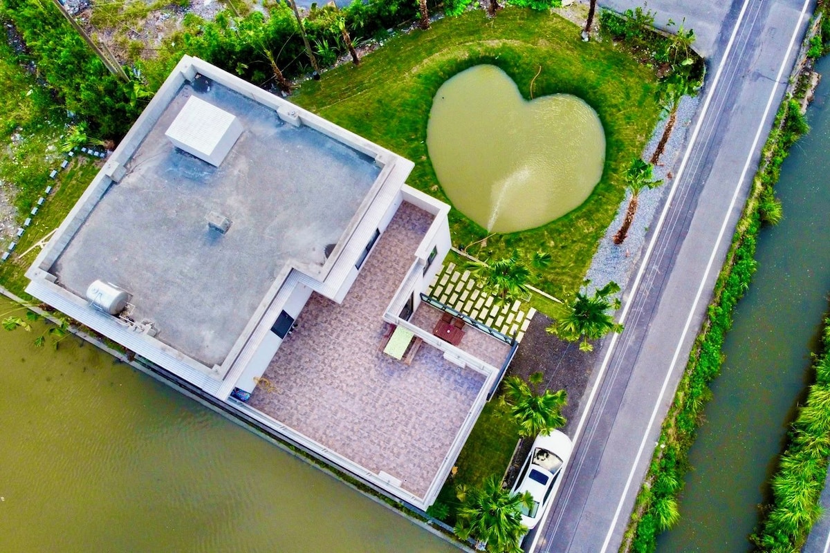 An aerial view of a building with a rooftop terrace, a heart-shaped pond, and a road adjacent to a waterway.