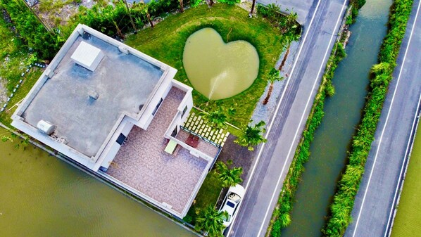An aerial view of a building with a rooftop terrace, a heart-shaped pond, and a road adjacent to a waterway.