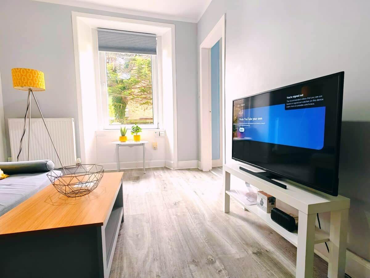A modern living room with a flat-screen TV, a wooden coffee table, a floor lamp, and a window with blinds.